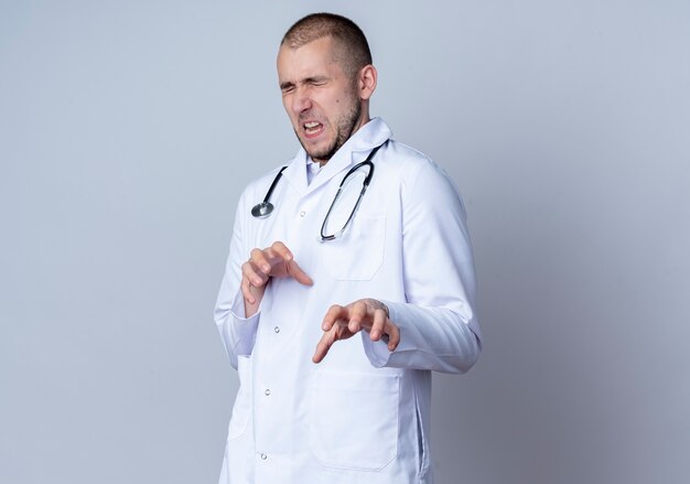 Geërgerd jonge mannelijke arts die medische mantel en stethoscoop om zijn nek draagt die hand met gesloten ogen uitrekt die op wit met exemplaarruimte wordt geïsoleerd
