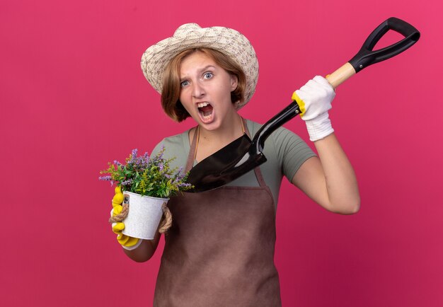 Geërgerd jonge Slavische vrouwelijke tuinman die tuinierende hoed en handschoenen draagt die bloemen in bloempot en schop op roze houden