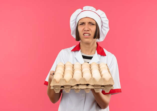 Geërgerd jonge vrouwelijke kok in het karton van de chef-kok uniforme bedrijf eieren geïsoleerd op roze met kopie ruimte