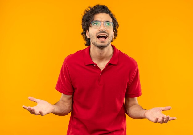 Geërgerd jongeman in rood shirt met optische bril houdt handen open geïsoleerd op oranje muur