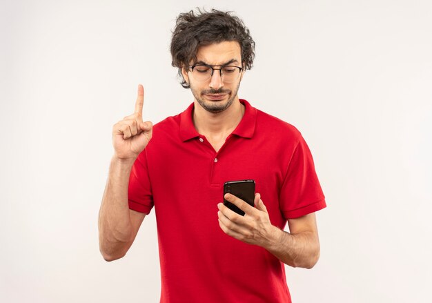 Geërgerd jongeman in rood shirt met optische bril houdt telefoon vast en wijst omhoog geïsoleerd op een witte muur