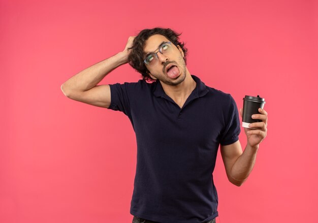 Geërgerd jongeman in zwart shirt met optische bril, steekt tong uit en houdt koffiekopje geïsoleerd op roze muur