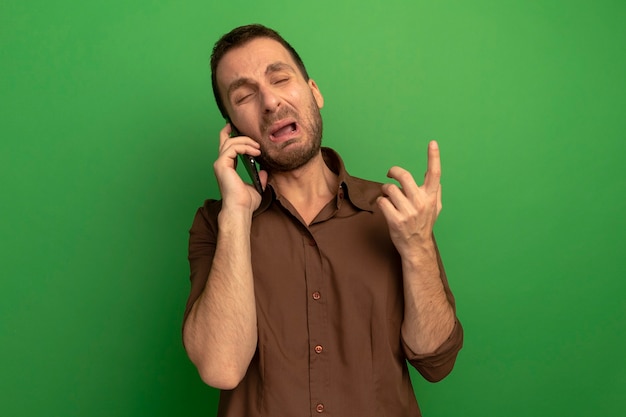 Geërgerd jongeman praten over telefoon met gesloten ogen geïsoleerd op groene muur