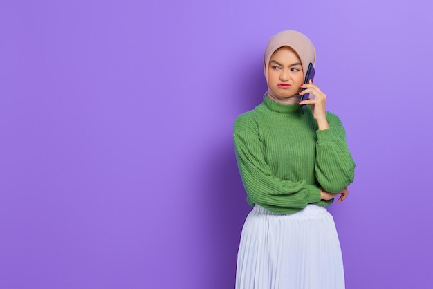 Geërgerd mooie aziatische vrouw in groene trui en hijab praten op een mobiele telefoon terwijl ze opzij kijkt geïsoleerd over paarse achtergrond