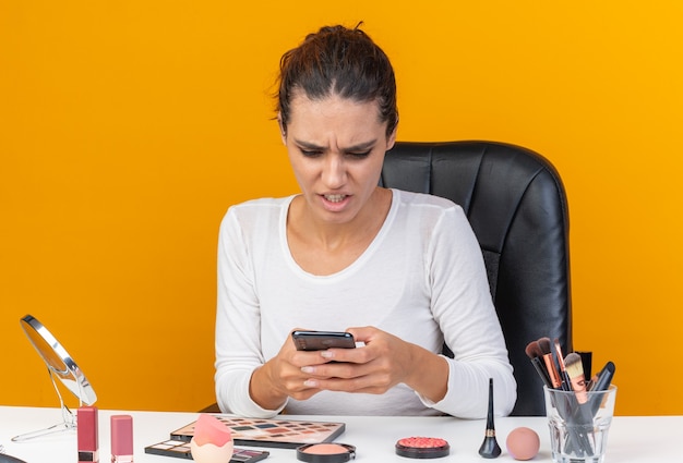 Geërgerd mooie blanke vrouw zittend aan tafel met make-up tools houden en kijken naar telefoon geïsoleerd op oranje muur met kopieerruimte