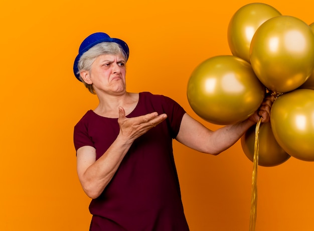 Geërgerd oudere vrouw met feestmuts houdt en wijst naar helium ballonnen geïsoleerd op oranje muur met kopie ruimte