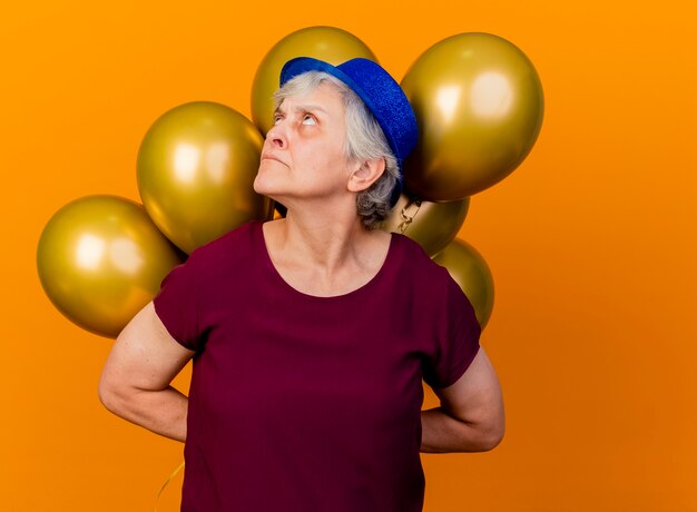 Geërgerd oudere vrouw met feestmuts houdt helium ballonnen achter opzoeken op oranje