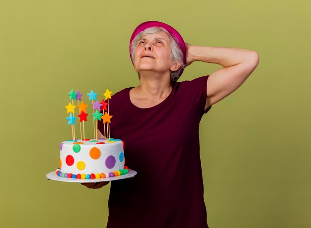 Geërgerd oudere vrouw met feestmuts legt hand op het hoofd achter het houden van verjaardagstaart geïsoleerd op olijfgroene muur met kopie ruimte