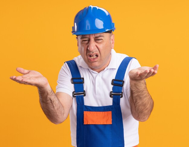 Geërgerd volwassen bouwer man in uniform houdt handen open geïsoleerd op oranje muur