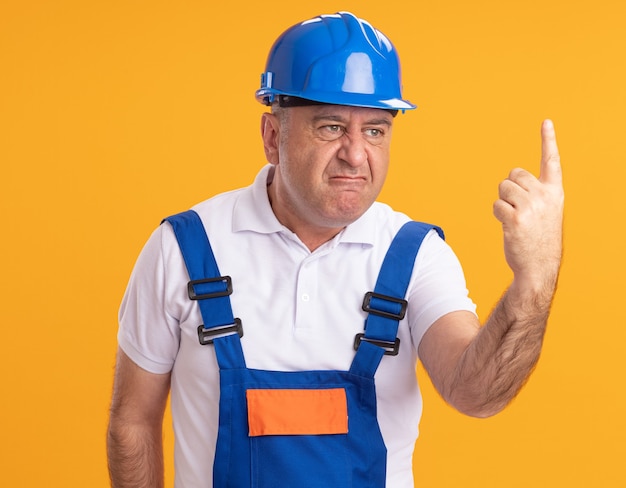 Geërgerd volwassen bouwer man in uniform toont wijsvinger geïsoleerd op oranje muur