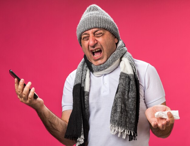 Geërgerd volwassen zieke blanke man met sjaal om nek dragen winter hoed met telefoon en servet geïsoleerd op roze muur met kopie ruimte