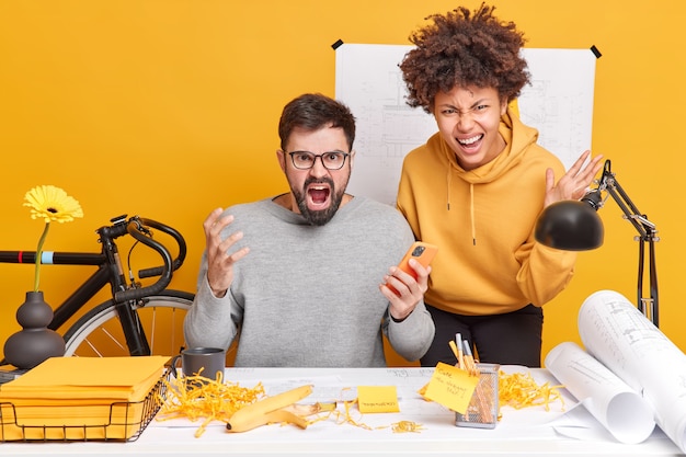 Geërgerde boze collega's hebben woedende gezichtsuitdrukkingen poseren op kantoor op desktop schreeuwen luid genoeg van veel werk, gekleed terloops gekleed verslag opstellen tekenen blauwdrukken