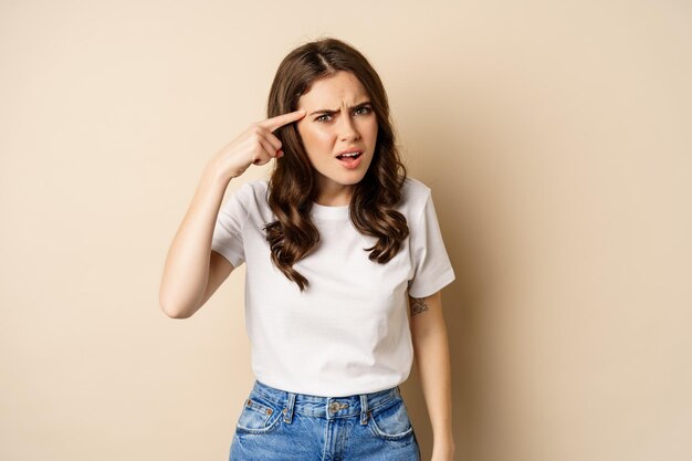 Geërgerde en boze vrouw die met de vinger naar het hoofd wijst, iemand uitscheldt, over een beige achtergrond staat