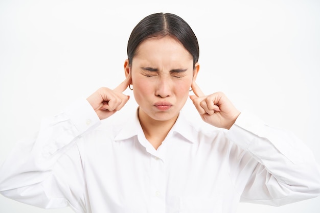 Gratis foto geërgerde jonge aziatische bedrijfsmedewerker dame schreeuwt en sluit haar oren met gefrustreerde vingers