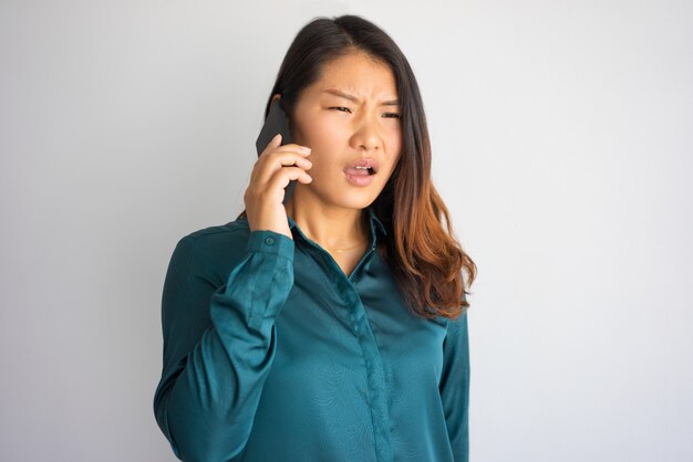 Geërgerde jonge Aziatische vrouw in vrijetijdskleding die op telefoon spreekt.