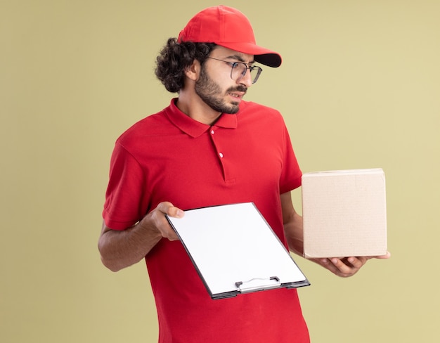 Geërgerde jonge blanke bezorger in rood uniform en pet met een bril met cardbox en klembord kijkend naar cardbox