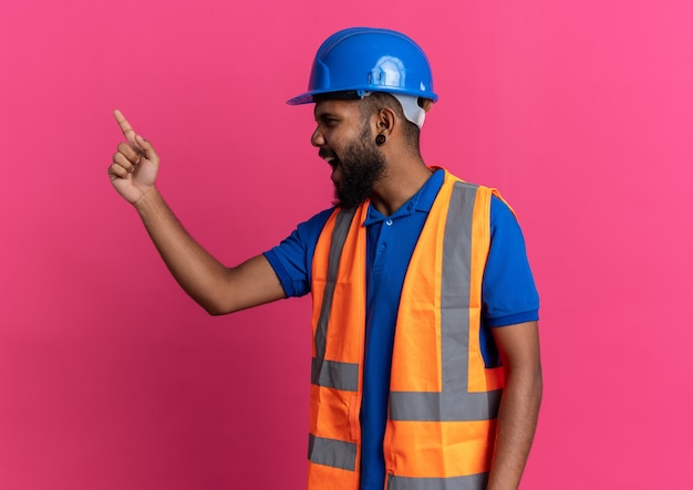 geërgerde jonge bouwman in uniform met veiligheidshelm schreeuwend tegen iemand die naar kant kijkt geïsoleerd op roze muur met kopieerruimte