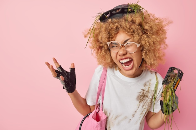 Geërgerde jonge gekrulde vrouw leidt actieve levensstijlgebaren en schreeuwt boos moet stoppen vanwege fietspech draagt vuil t-shirt houdt pedaal met gras vast en probeert haar transport te repareren