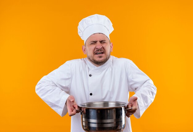Geërgerde jonge knappe kok in uniform van de chef-kok met ketel met gesloten ogen op geïsoleerde oranje muur