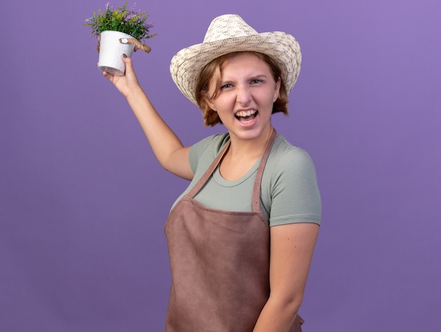 Geërgerde jonge slavische vrouwelijke tuinman met een tuinhoed met bloemen in een bloempot geïsoleerd op een paarse muur met kopieerruimte