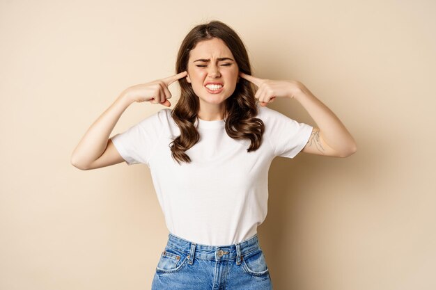 Geërgerde jonge vrouw sloot de oren van hard geluid, voelde zich ongemakkelijk en klaagde, staande tegen een beige achtergrond.