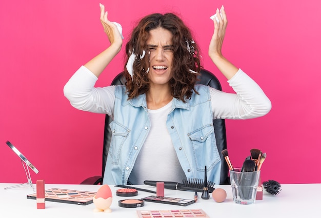 Geërgerde mooie blanke vrouw die aan tafel zit met make-uptools die haarmousse toepassen op een roze muur met kopieerruimte