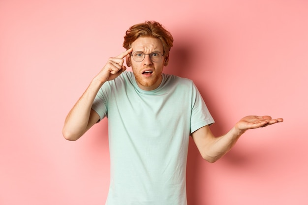 Geërgerde roodharige man die iemand uitscheldt die met de vinger naar het hoofd wijst en zijn schouders ophaalt over roze...