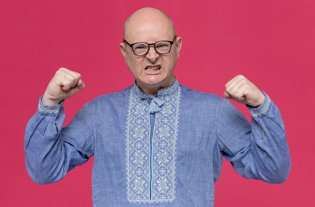 Geërgerde volwassen Slavische man in blauw shirt met een bril die vuisten houdt