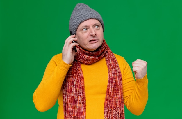 Geërgerde volwassen slavische man met wintermuts en sjaal om zijn nek die aan de telefoon praat en de vuist naar de zijkant kijkt