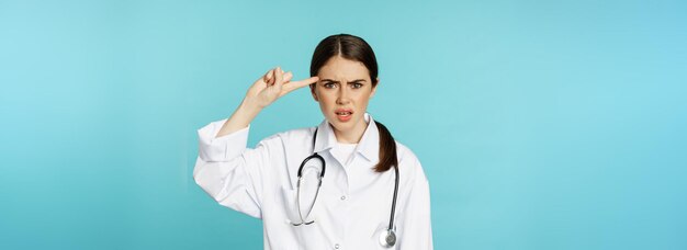 Geërgerde vrouwelijke arts die met de vinger naar het hoofd wijst en iemand een uitbrander geeft, gek of vreemd die voorover staat?