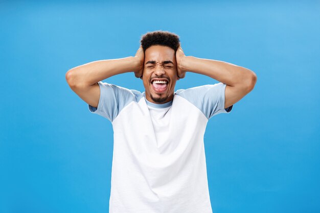 Geest waait uit veel informatie. Portret van een verontruste Afro-Amerikaanse mannelijke student in een t-shirt die handen op het hoofd houdt en met gesloten ogen schreeuwt om boos te worden over de blauwe muur