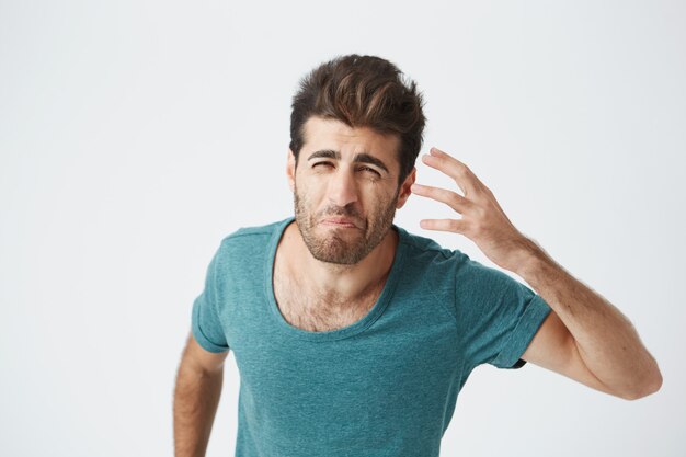 Gefrustreerd aantrekkelijke bebaarde blanke man in trendy blauwe tshirt, met knorrige gekke uitdrukking gebarend met de hand. Menselijke gezichtsuitdrukkingen en emoties.