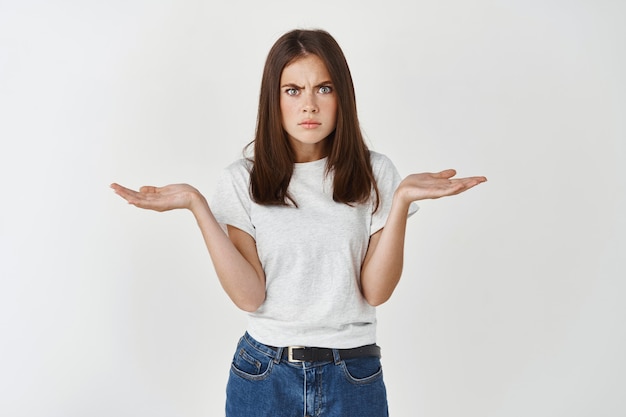 Gefrustreerd en verward, brunette vriendin die ruzie heeft, vragen heeft, teleurstelling uit, schouderophalend met de handen zijwaarts gespreid in ontzetting, besluiteloos en aarzelend, witte muur.