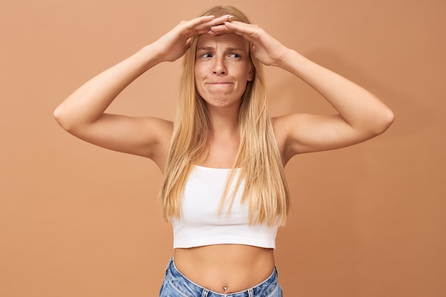 Gefrustreerde angstige jonge vrouw met doordringende hand in hand op haar hoofd en het nastreven van lippen poseren geïsoleerd met verbaasde blik