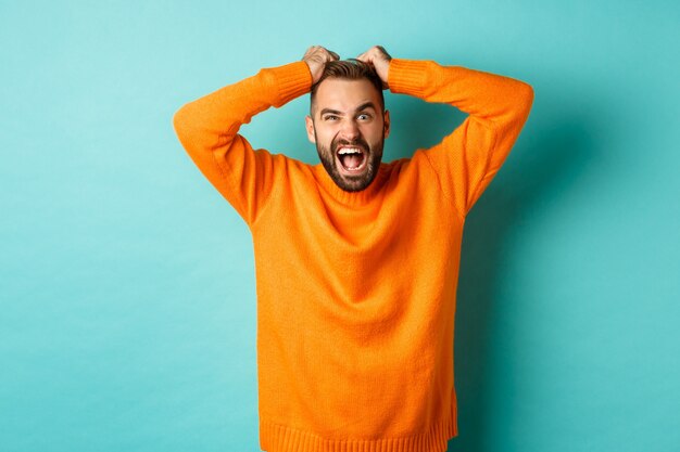 Gefrustreerde man die schreeuwde, haar haar uittrekt en boos schreeuwt, zijn geduld verliest en er boos uitziet, staande over de lichtblauwe muur.