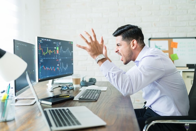 Gefrustreerde Spaanse jonge freelance aandelenhandelaar die schreeuwt terwijl hij naar grafieken op het computerscherm kijkt