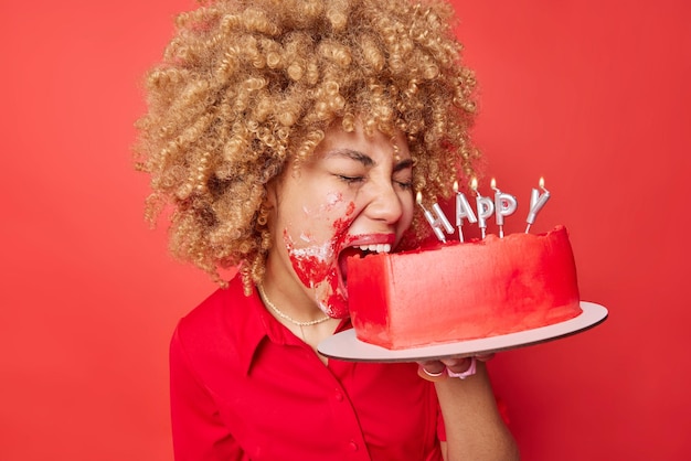 Gefrustreerde vrouw met krullend haar bijt cake besmeurd met room viert valentijnsdag alleen heeft slecht depressieve stemming poses tegen levendige rode achtergrond verwende vakantie en eenzaam viering concept