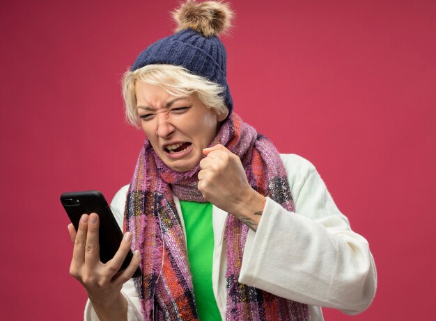 Gefrustreerde zieke ongezonde vrouw met kort haar in warme sjaal en muts die zich onwel voelen met smartphone gebalde vuist staande over roze achtergrond