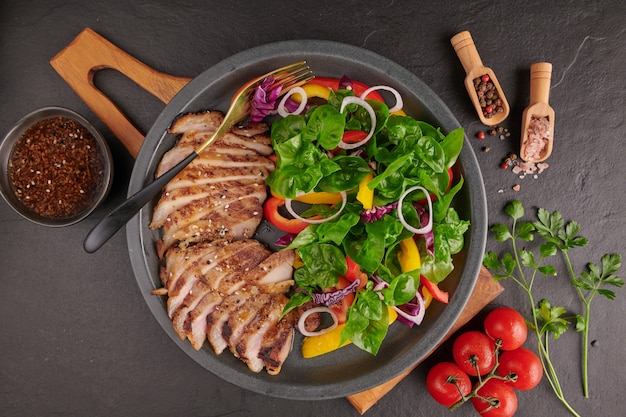 Gegrilde steak met verse groente, paprika, tomaten, rode ui, roze peper en kruiden. Zelfgemaakt lekker eten. Concept voor lekkere en gezonde maaltijd. Zwart stenen oppervlak. Varkenslapje vlees met salade