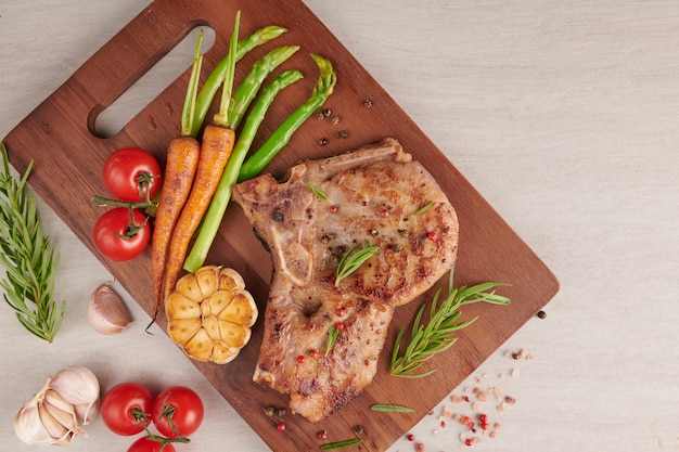 Gegrilde varkenssteak van een zomerse BBQ geserveerd met groenten, asperges, worteltjes, verse tomaten en kruiden. Gegrilde biefstuk op houten snijplank op stenen oppervlak. bovenaanzicht.