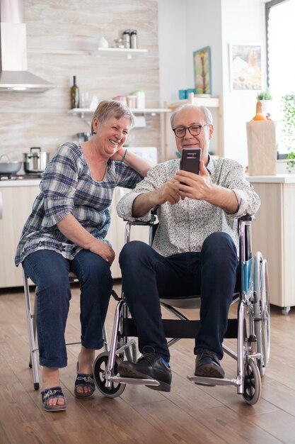 Gehandicapte senior man in rolstoel en zijn vrouw lachen en browsen op moderne smartphone in de keuken. Verlamde oude man en zijn vrouw hebben een online conferentie.