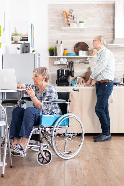 Gehandicapte senior vrouw in rolstoel die vanuit huis werkt op laptop in de keuken en man die ontbijt bereidt. gehandicapte zakenvrouw, gehandicapte ondernemer verlamming voor oudere gepensioneerde vrouw