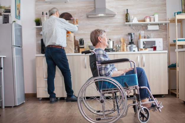 Gratis foto gehandicapte senior vrouw zit in een rolstoel in de keuken en kijkt door het raam. leven met gehandicapte. echtgenoot helpt vrouw met een handicap. bejaard paar met gelukkig huwelijk.