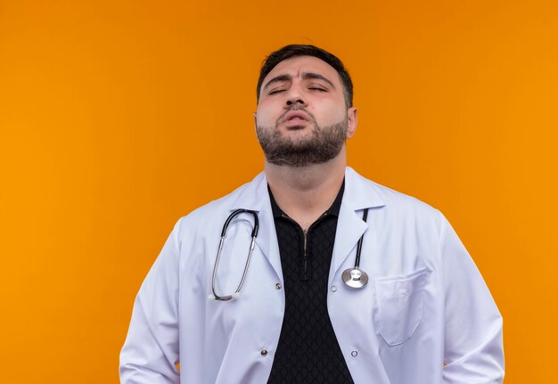 Gehinderd jonge bebaarde mannelijke arts dragen witte jas met stethoscoop op zoek moe nad benadrukt sluiten ogen