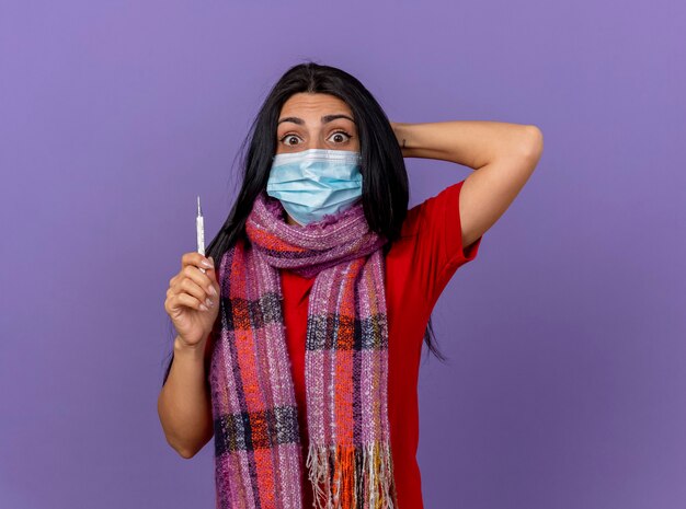 Geïmponeerd jong Kaukasisch ziek meisje dat masker en sjaal draagt die thermometer houdt die hand achter hoofd houdt dat op purpere muur met exemplaarruimte wordt geïsoleerd
