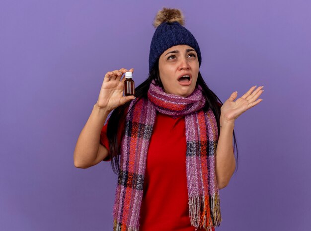 Geïmponeerd jong Kaukasisch ziek meisje dat winter muts en sjaal draagt en medicijn in glas houdt opzoeken met lege hand geïsoleerd op paarse muur met kopie ruimte
