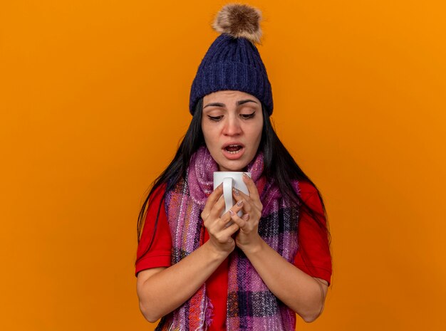 Geïmponeerd jong Kaukasisch ziek meisje die de winterhoed en sjaal dragen die kop thee houden die binnenkop kijken die op oranje achtergrond met exemplaarruimte wordt geïsoleerd