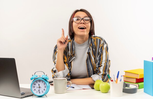 Geïmponeerd jong studentenmeisje die glazen dragen die bij bureau zitten dat vinger opheft die op wit wordt geïsoleerd