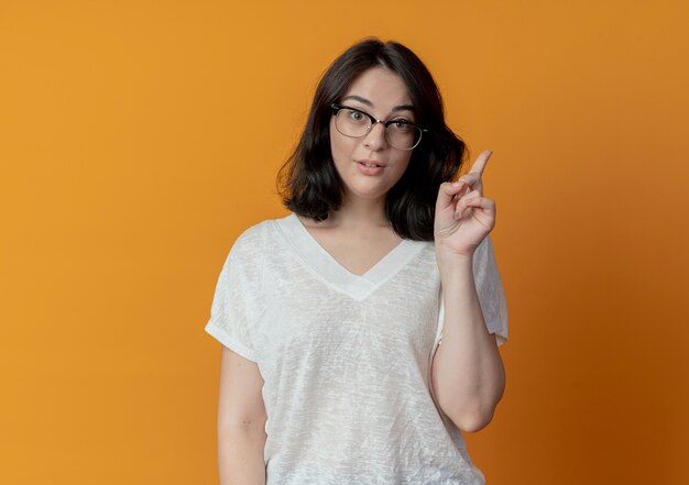 Geïmponeerd jong vrij Kaukasisch meisje dat glazen draagt die vinger opheffen die op oranje achtergrond met exemplaarruimte wordt geïsoleerd