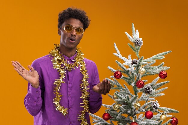 Geïmponeerde jonge Afro-Amerikaanse man met bril met klatergoud slinger rond de nek staande in de buurt van versierde kerstboom op oranje achtergrond